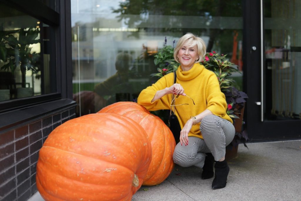 Deborah Boland kneeling beside giant pumpkins wearing gold oversized Turtleneck and check cropped pants