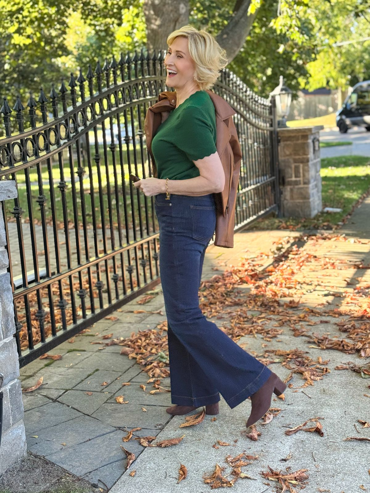 Deborah wearing a green scalloped short-sleeve top and dark high-waisted bootcut jeans with a brown belt, smiling with arms raised among autumn leaves