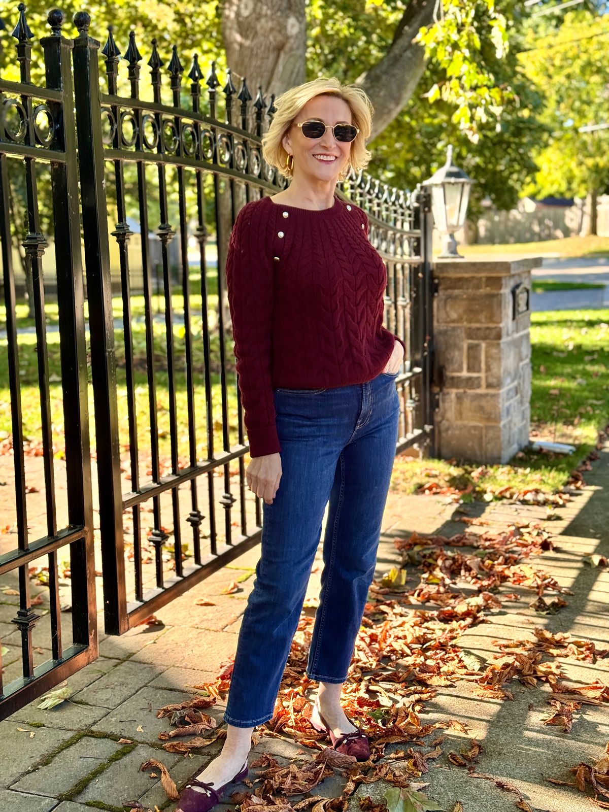 Deborah wearing a burgundy cable knit sweater with gold button details and dark wash straight-leg jeans standing near a black iron fence surrounded by fall leaves