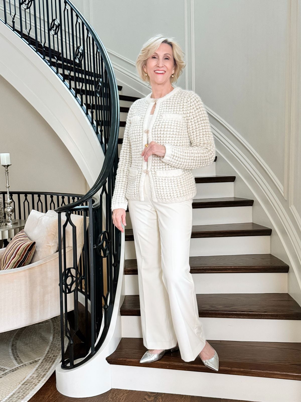 Deborah wearing a cream tweed jacket, white pants, and pointed silver pumps while standing on a staircase.