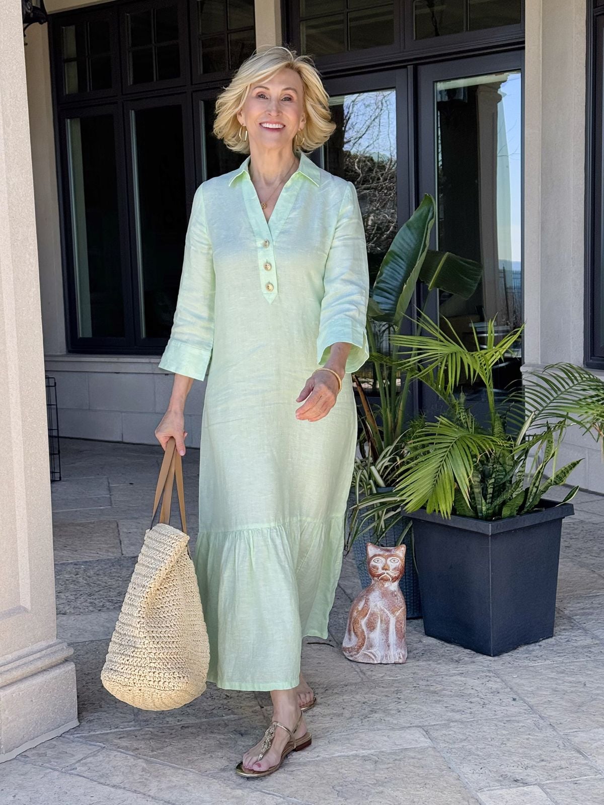 Deborah Boland modeling a tiered sage green linen maxi dress with three quarter length sleeves and a relaxed fit styled with a straw tote bag.