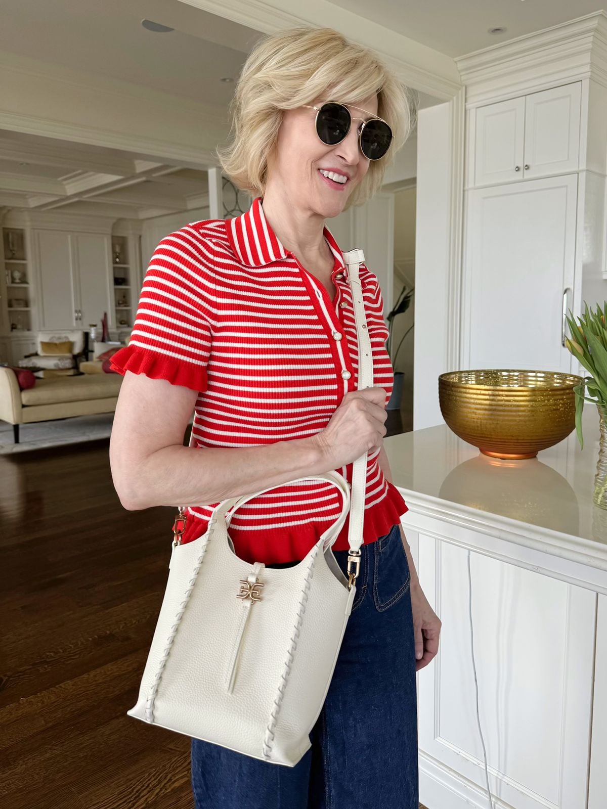Deborah wearing a structured white leather bucket bag with braided edge details and gold hardware, styled with a red and white striped polo shirt and denim.