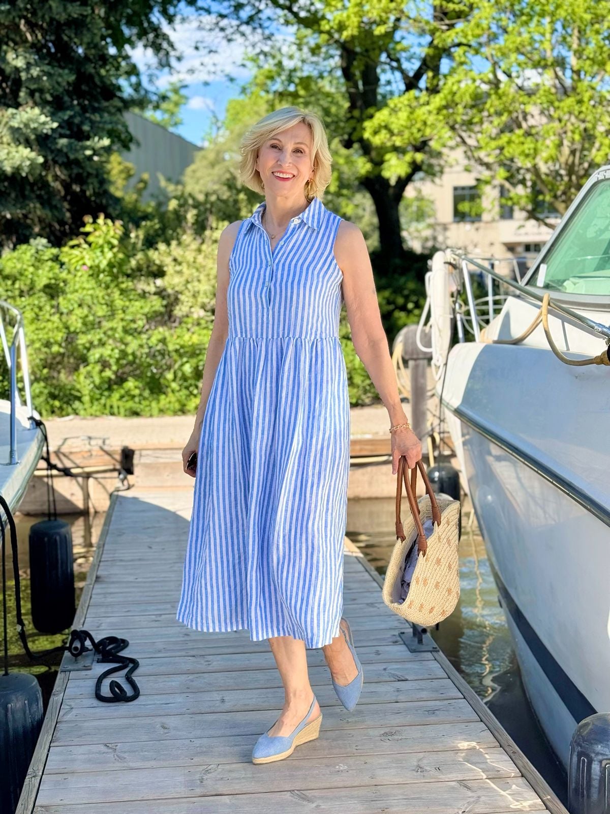 Deborah Boland wearing a blue and white striped linen midi dress with a woven tote and espadrilles in a modern coastal style outfit.