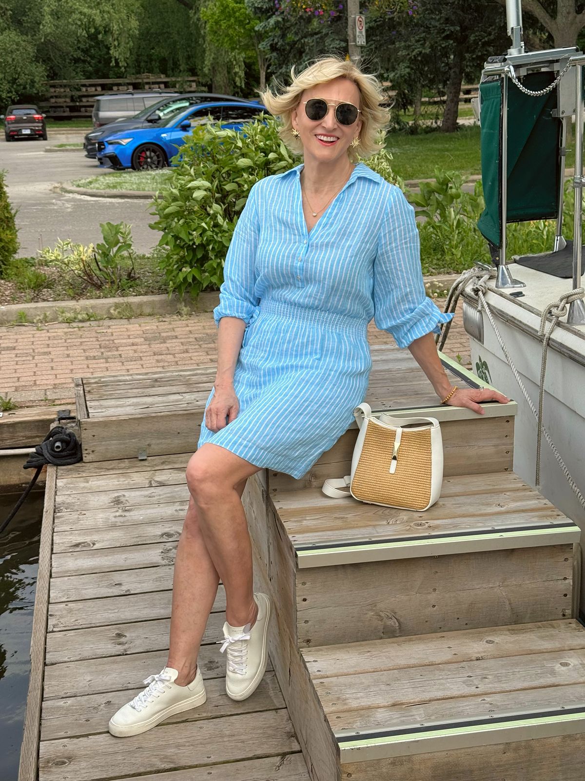 Deborah Boland wearing a blue striped linen dress with white leather sneakers and a woven crossbody bag for a classic coastal style outfit.
