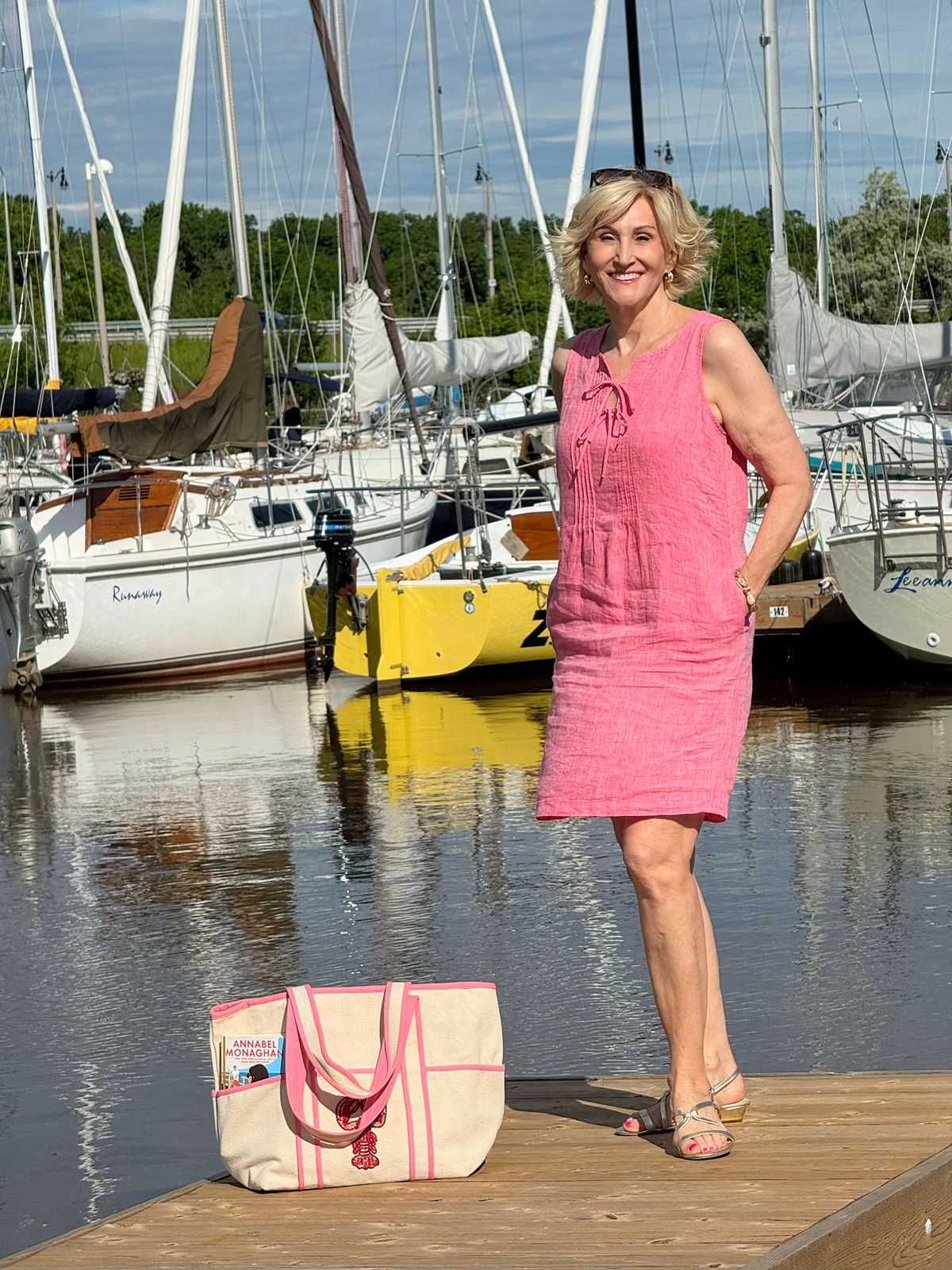 Deborah Boland wearing a coral pink linen shift dress with metallic sandals and a canvas tote bag in a modern coastal style outfit at a marina.