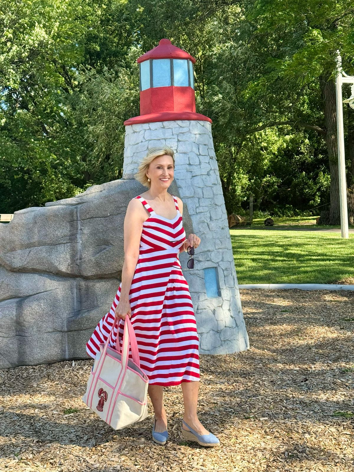 Deborah Boland wearing a red and white striped midi dress with espadrilles and a canvas tote in a classic nautical style outfit by a lighthouse.