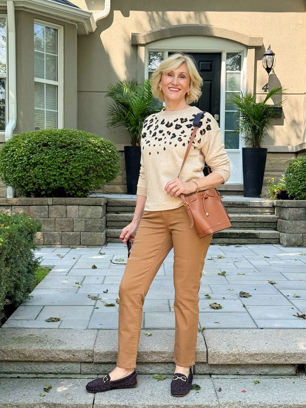 Deborah wearing a leopard sweater, brown leather bag, brown straight leg pants and leopard loafers.